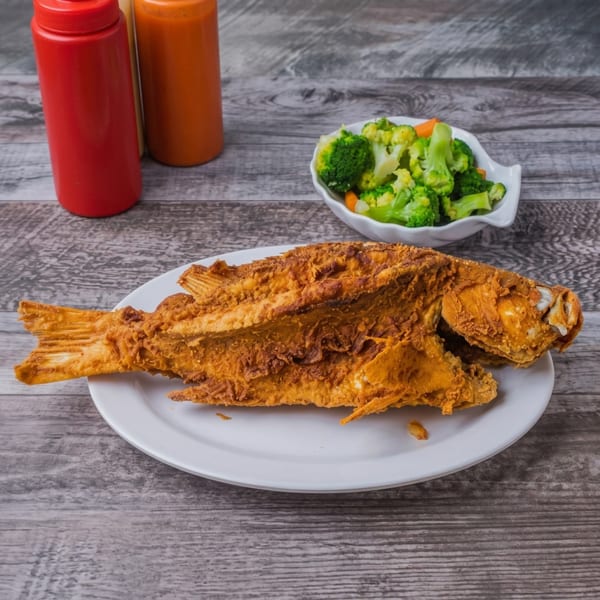 Whole fried fish on a white plate with a side of broccoli and carrots, plus two sauce bottles.