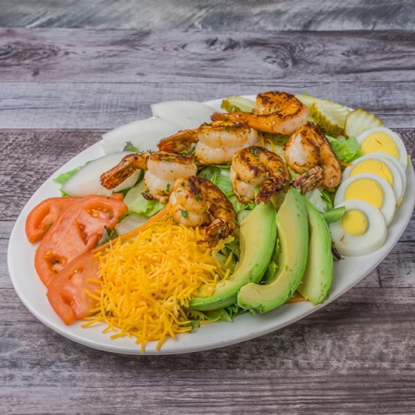 Grilled shrimp salad with lettuce, tomatoes, cheese, avocado, eggs, and pickles on a white plate.